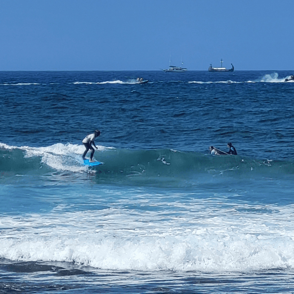 A surfer on the water in Las Americas tenerife. There are a couple of big waves and on the horizon you can see 2 boats.